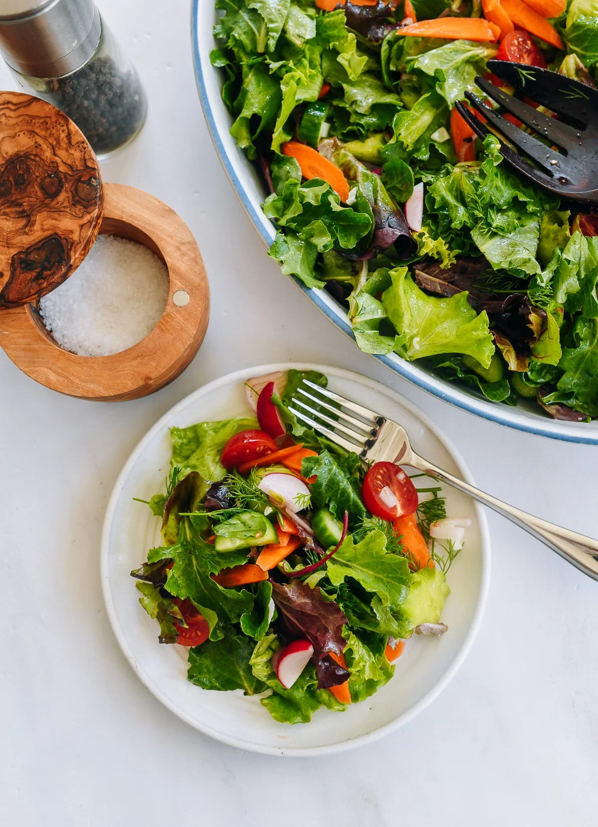 Two bowls of fresh side salad on a marble counter with serving spoons