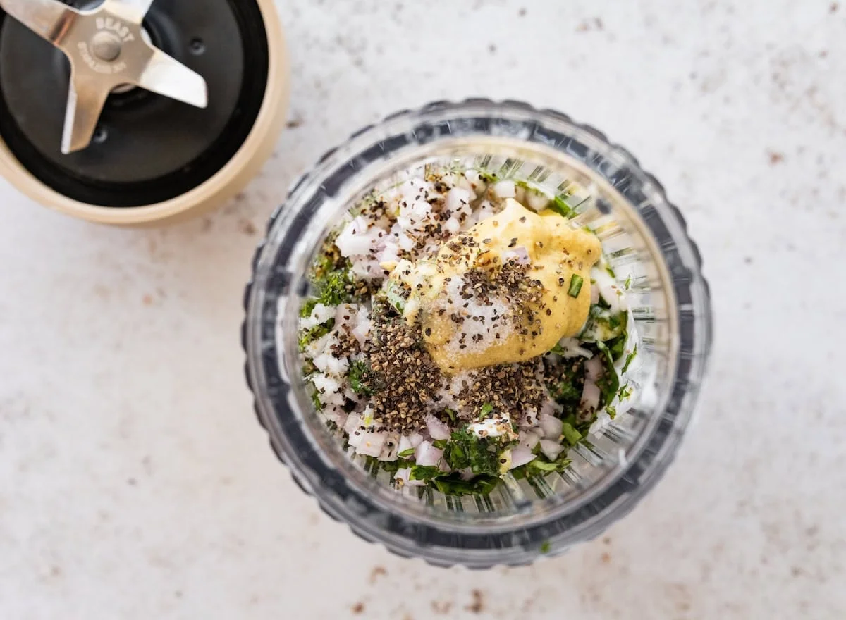Blender cup filled with herbs, shallot, and mustard before blending