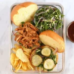 BBQ pulled chicken sandwich with slaw, pickles, and chips on a tray