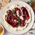 Layered goat cheese dip with raspberry jam, hot honey, rosemary, and red pepper flakes on a platter with crackers