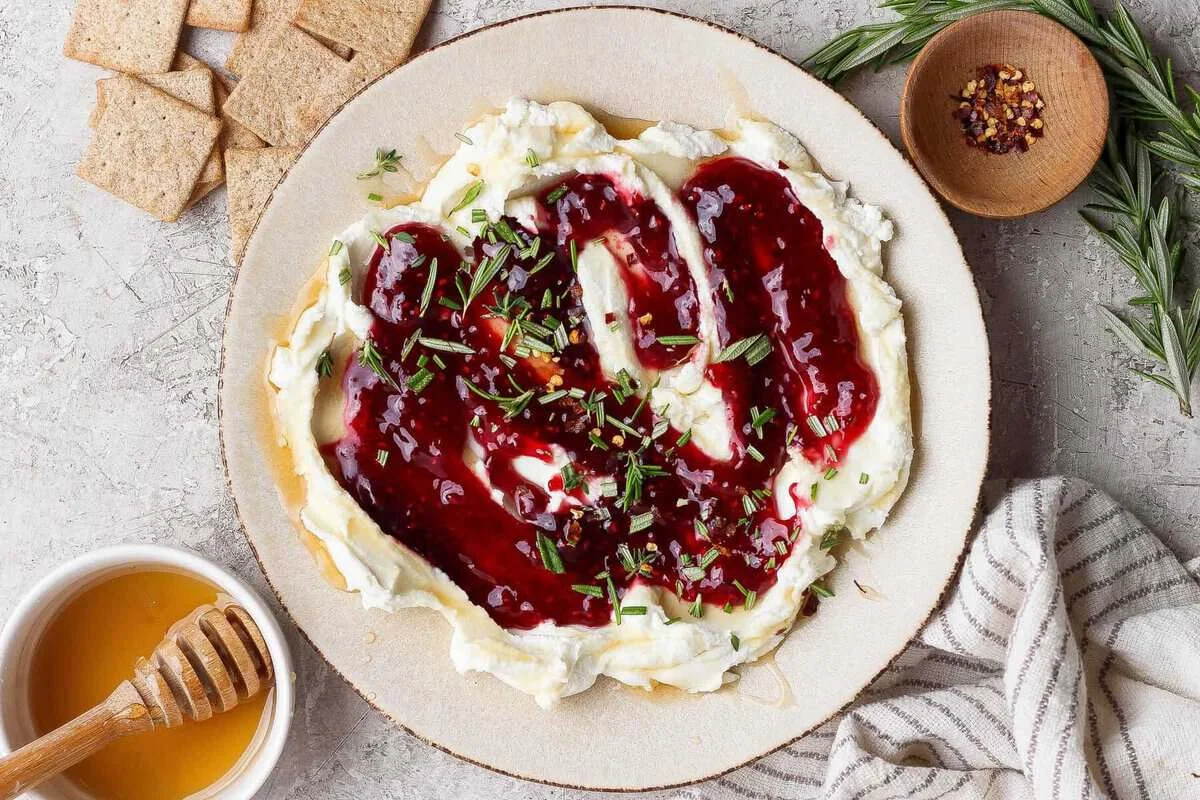 Layered goat cheese dip with raspberry jam, hot honey, rosemary, and red pepper flakes on a platter with crackers